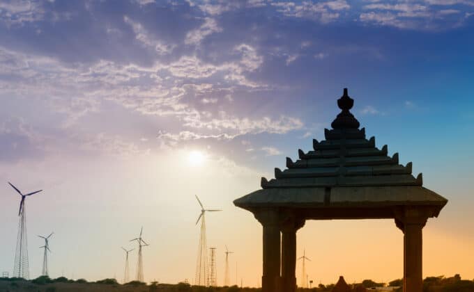 A chhatri (domed pavilion) stands in silhouette against a blue and orange sunset. Modern wind turbines are in the distance.