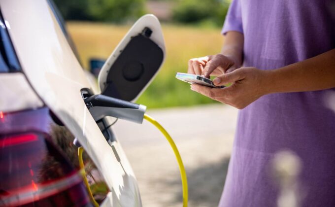 woman in purple shirt stands next to charging electric vehicle using smart phone app